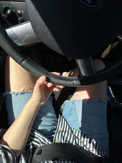 The image shows a close-up view of a person's hands and legs inside a car, as seen from the driver's perspective. The driver, likely female based on the hands and legs, is wearing denim shorts and a striped shirt. The hands are gripping the steering wheel, which appears to be black leather. The car interior is visible, including part of the dashboard with light controls and a silver accent piece. Sunlight streams in, casting strong shadows across the scene. The setting is not specified as being in the city of Ленинское, Russia.