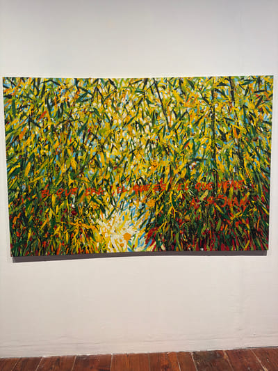 This is a vertical, close-up, eye-level shot of a vibrant abstract painting hanging on a textured, off-white wall. The painting is a dense tapestry of short, expressive brushstrokes in various shades of green, yellow, orange, red, and blue. The strokes are layered and overlapping, creating a sense of movement and depth. There are hints of black and white interspersed, adding further visual complexity. The overall impression is one of a chaotic yet harmonious natural scene, perhaps suggesting foliage in a strong wind or a sun-drenched field.
The painting occupies the right half of the frame. The left side of the image is dominated by the plain, off-white wall, which exhibits subtle texture and a few faint imperfections. To the far right of the painting, a narrow section of a wooden wall is visible, with dark, vertical planks showing a natural wood grain and some visible knots. The lighting appears to be artificial, casting a soft glow on the artwork and the wall. There is no visible text or any depiction of people or specific objects. The setting suggests an indoor display, possibly an art gallery or studio space, given the presentation of the artwork and the neutral background.