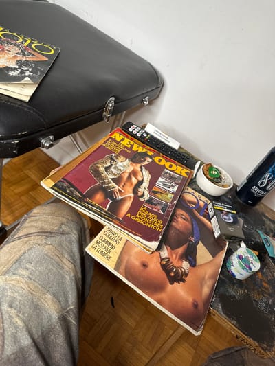 The image features a cluttered tabletop with various objects. A black guitar case sits to the left, with a magazine partially visible. Several magazines, including "New Look" and "OTO Magazine" are stacked, featuring images of scantily clad people. Other items include a remote control, a small white dish with a green object, a can of "80" energy drink, a pack of cigarettes, and a decorative container. The tabletop has a dark, mottled surface, and the overall scene appears disorganized. A person's legs and pants are also visible.