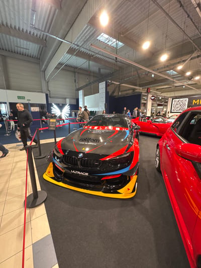 The image displays an indoor automotive exhibition at a venue in Wolica, Poland. In the foreground, a heavily modified black BMW M4 race car with carbon fiber elements, accented with bright red, blue, and yellow aerodynamic components and "rotiform" and "ULTRACE" branding, is prominently displayed. Japanese text is visible on the upper windshield banner. The vehicle is situated on a dark grey carpeted area, separated from a light-tiled walkway by red stanchions and a red rope barrier.

To the immediate right of the BMW, the front-left portion of a glossy red vehicle is partially visible. Further back and to the right, a red convertible sports car is parked. Multiple individuals are present throughout the exhibition space; one man in a dark hoodie walks past the roped-off area on the left, while other attendees in casual attire are scattered, observing the vehicles and congregating in the background.

The background includes various exhibition elements: an illuminated white, wing-shaped art installation is visible to the left of the BMW. A large black and white QR code banner hangs on a wall section in the upper right. The ceiling is high, industrial-style, with exposed beams, ventilation ducts, fluorescent lighting fixtures, and several skylights allowing natural light. A white "WYJŚCIE" (EXIT) sign in Polish is mounted above a doorway on the far left wall.