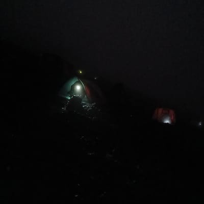 The scene depicts a high-altitude campsite at night near Horombo Huts, Tanzania. The environment is dark and appears misty, causing visible light sources to diffuse. In the left-center foreground, a two-tone tent, primarily green and red, is illuminated by a strong white light source positioned at its open entrance. An indistinct, dark figure is discernible near this light source. The immediate ground surrounding this tent is uneven, dark, and appears to be a wet or rocky natural surface. To the mid-right, a second tent, reddish-orange in hue, glows faintly from an internal light source. Multiple other small, diffused light points are scattered in the dark background, indicating additional tents or camp activity.
