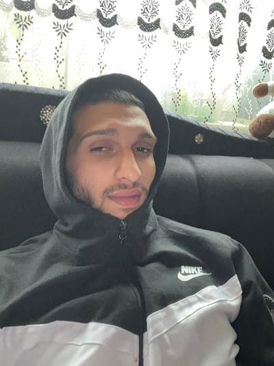 A man wearing a gray Nike hoodie is lying on a dark gray couch. The background features a white, patterned curtain and potentially a window. The location in Giengen an der Brenz, Germany, cannot be determined from the image.