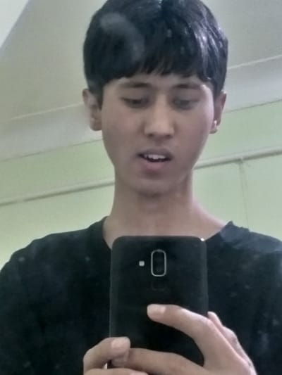 The image is a mirror selfie, originally rotated 90 degrees counter-clockwise. When viewed upright, it shows a young person taking a photo of their reflection.
The subject is a young individual, likely a male teenager or young adult, positioned in the upper center of the frame. They have short, dark hair that appears wet or damp, falling over their forehead in a fringe. The hair has noticeable wet strands and reflections, suggesting they may have recently washed it. The person has a light complexion, with dark eyes that are partially closed or squinting. Their mouth is slightly open, revealing some teeth, and their expression is neutral or slightly pensive.
They are wearing a dark-colored (likely black) short-sleeved t-shirt, which is visible on their upper torso.
The activity is clearly a mirror selfie. The person's right hand is visible on the right side of the frame, holding a black smartphone. The back of the phone is facing the viewer (reflected in the mirror), revealing a vertical module on the upper center, which appears to house dual cameras or a single camera with a flash/sensor, and a distinct, rounded rectangular component below it, likely a fingerprint sensor.
The background behind the person in the reflection is a plain, light-colored wall or surface, possibly off-white or a very light green. Faint vertical lines are visible on the left side of the background, suggesting a door frame or wall paneling. The setting appears to be indoors, likely a bathroom or bedroom. The lighting is uniform indoor lighting, contributing to the overall slightly muted tone of the image.