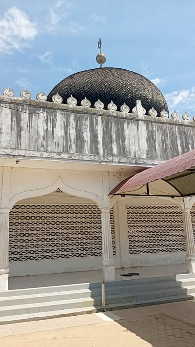 The image shows a mosque with a black, scale-covered dome topped by a crescent moon finial. The dome rests on a white structure with decorative elements along its upper edge. Below this, a white arched entryway with columns leads to an area with a patterned brick wall. Steps lead up to this area. To the right, a covered patio area has a reddish-brown metal roof. The ground in front of the steps is paved with sand and decorative tiles. The sky is blue with scattered white clouds. The location is the mosque in Alueanou Barat, Indonesia.