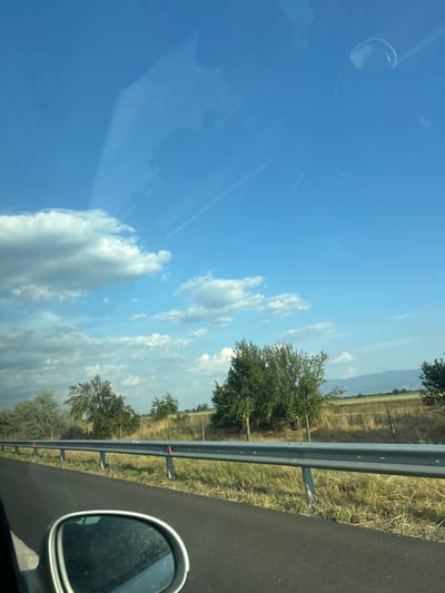 The image captures a roadside view from inside a vehicle, likely during daylight. The scene is dominated by a bright blue sky with scattered white clouds, suggesting fair weather. On the right side of the frame, a dark asphalt road is visible, bordered by a metal guardrail. Beyond the guardrail, a dry, grassy field stretches towards a line of trees and distant hills. The trees vary in size and foliage density, indicating a rural or semi-rural setting. A silver side-view mirror, possibly from the vehicle the photo was taken in, is prominently featured in the lower right corner. Reflections are visible on the car's window in the upper left quadrant. There is no visible text in the image. The perspective suggests the photo was taken while the vehicle was in motion. The time of day appears to be late morning or early afternoon, given the bright sunlight. The location is identified as Tsalapitsa, Bulgaria.