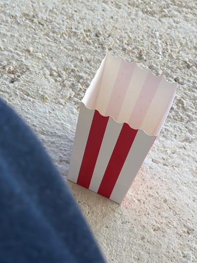 The image shows a close-up, high-angle shot of a red and white striped popcorn box resting on a textured, light-colored surface, likely concrete or pavement. A person's dark blue clothing is visible in the lower left corner of the frame, suggesting they are seated or leaning near the box. A small, thin object, possibly a piece of straw or grass, is visible on the ground to the left of the popcorn box. The background is a rough, granular texture of a light beige or gray color. The lighting suggests an outdoor environment, possibly in a public area like a park or street in Sant Vicenç de Montalt, Spain.