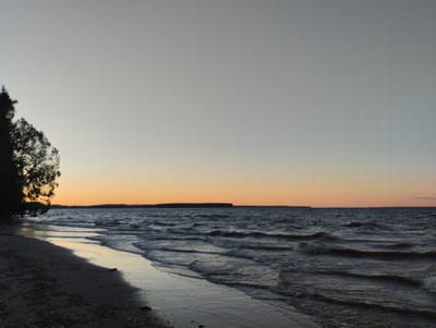 The image depicts a wide, calm lake at sunset. The sky is a pale gradient, transitioning from a soft grey at the top to a warm orange hue near the horizon. A distant, dark treeline borders the lake. In the foreground, a sandy beach curves into the frame, with gentle waves lapping at its edge. A silhouette of a tree with sparse branches is visible on the far left, partially obscuring the shoreline. The water is dark and reflects the muted colors of the sky, with small whitecaps indicating a light breeze. The overall scene is serene and natural, likely a natural landscape within or near Zavolzhye, Russia. There are no people or animals present in the image.