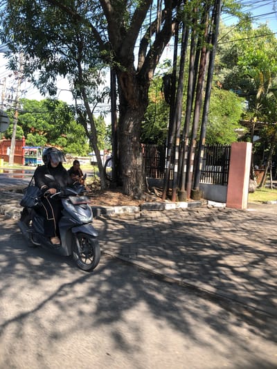A medium shot captures a woman riding a dark grey Honda scooter on a paved street in Buduran, Indonesia. She wears a black hijab, a black long-sleeved outfit with tan trim, a black helmet with a clear visor, and tan sandals. A black shoulder bag is slung over her left shoulder. The scooter has a license plate reading "4784 MCK". In the background, behind the scooter, another person is seated on the ground, appearing to be holding a phone. The scene is bordered by a lush green environment with trees and foliage, and electrical poles line the right side of the frame. A dark metal fence with a pink concrete pillar is visible further in the background. The ground is a mix of asphalt and patterned paving stones, and dappled sunlight casts tree shadows across the road. A red and green building with a blue roof is visible on the left side of the street, and a street cart is parked in front of it.