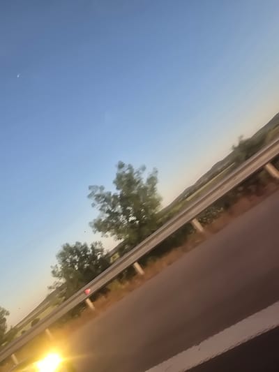 The image shows a view from a vehicle, likely a car or train. The sky is blue and mostly clear. Along the top of the image, there is a road with vegetation. The right side of the image shows part of the vehicle interior, a brown surface with a white edge. There is no discernible location of Yazdach, Bulgaria.