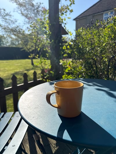 This media file captures an outdoor garden scene, likely a backyard, under bright, sunny conditions. The primary subjects are a light brown or tan ceramic mug resting on a round, blue metal garden table. The table exhibits a slightly textured or speckled surface. To the right of the table, a part of a matching blue metal garden chair is visible, showing its slatted design.

The background is rich with natural elements. Lush green foliage, comprising trees and bushes, dominates the left and central portions, rendered with a pleasant bokeh effect due to a shallow depth of field. Some small red or pink blossoms can be discerned within the greenery. Beyond the vegetation and slightly to the left, a portion of a house with a dark roof and a light-colored wall with a window is visible against a clear blue sky. A wooden fence runs along the middle ground, separating the garden area. A well-maintained green lawn is visible behind the fence and to the right.

The strong, clear sunlight casts distinct, elongated shadows from the mug, the table, and the surrounding foliage, indicating either early morning or late afternoon. The overall impression is one of peace and relaxation, typical of enjoying a beverage outdoors in a domestic garden setting in Hitchin, United Kingdom. There is no discernible activity taking place beyond the still life, and no visible text.