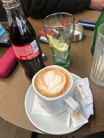 The scene depicts a close-up, overhead view of a table in an establishment located in Kumanovo, North Macedonia. Centrally positioned is a white ceramic cup containing a cappuccino adorned with heart-shaped latte art, resting on a matching saucer. On the saucer, two sealed white sugar packets, one featuring the brand "@bartrgovski", and a small white plastic spoon are present. To the left of the cup, a partially consumed clear glass Coca-Cola bottle stands upright, displaying a red label with Cyrillic text, including product information in Macedonian. A dark pink rectangular electronic device is visible to the left of the Coca-Cola bottle. To the right of the cappuccino cup, a green-tinted glass, embossed with a Coca-Cola logo, contains water with two lemon slices. Further right and slightly behind, an empty metallic ashtray is visible, followed by a tall, clear glass. The items are arranged on a light brown wooden table surface. Blurred dark clothing, indicative of at least two individuals, is visible in the background, confirming the presence of people at the table. No specific actions or interactions between individuals are discernible.
