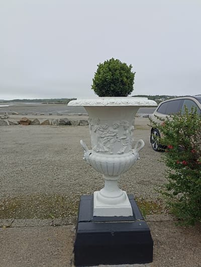 A white, ornate urn containing a round, green bush sits on a black pedestal in the foreground. The urn features relief carvings of figures. To the right of the urn, a light-colored SUV is parked. Behind the urn and SUV, a gravel area extends towards a body of water, likely a tidal estuary or sea, with a low, distant shoreline visible under a cloudy sky. The scene is located in Annagry, County Donegal, Ireland, a coastal area known for its rugged landscapes.