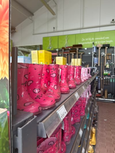 The image shows a retail display of pink children's rain boots with heart and star patterns, arranged on shelves in a store. The boots are stacked in rows, with the front row being the most prominent. Behind the boots, various gardening tools like rakes and brooms are visible on a dark wall. There are also yellow boxes stacked on a shelf. A price tag with the text "Kindermom. Weg PVS. Nu maar 28,50" is attached to one of the shelves. The store appears to be a garden center. The image was taken inside a store located in Tuk, Netherlands, which is evident from the text on the price tag referencing "Groenrijk," a Dutch garden center chain.