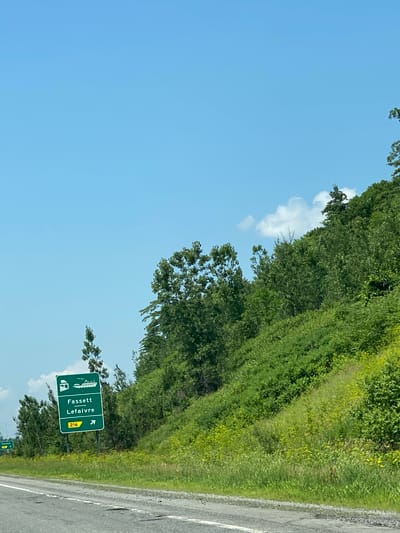 An exterior, eye-level, medium shot captures a bright, sunny day with a clear blue sky dominating the upper half of the frame. A verdant, tree-covered hill slopes upwards and to the right. In the foreground, a paved road runs horizontally across the bottom, with a white line marking its edge. To the left of the road, a green directional sign stands on a pole. The sign displays an image of a truck and a boat, followed by the text "Fassett Leflavre" and the number "216" within a yellow rectangle. An arrow pointing to the right is also visible on the sign. In the distance, a smaller green sign with "Fassett 216" is visible. The scene is set in Fassett, Canada, a municipality located in the Montérégie region of Quebec. The environment appears to be rural or semi-rural, with abundant vegetation and a highway-like road. No people are visible in the image.