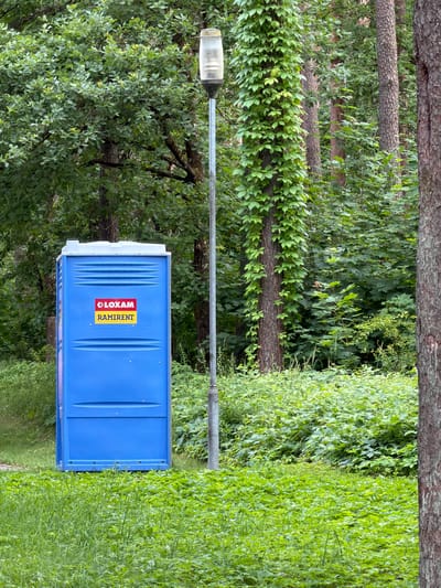A blue portable toilet is situated on a grassy area in front of a wooded backdrop. The toilet has two decals on its front: a red and white decal that reads "LOXAM" and a yellow and black decal that reads "RAMIRENT." A street lamp stands to the right of the toilet, its pole entwined with ivy. The area is surrounded by trees and dense green foliage. The scene appears to be in the city of Upeslejas, Latvia.