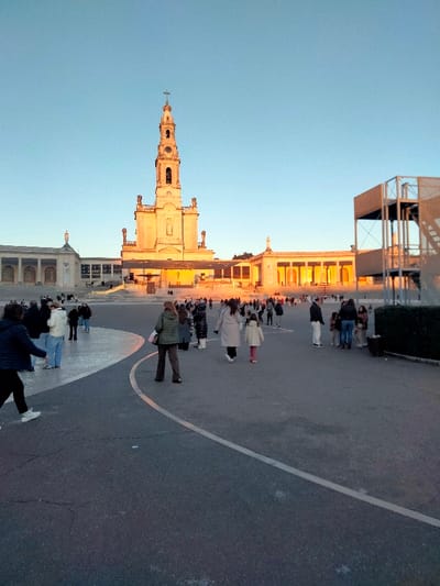 This image depicts a wide view of the Sanctuary of Fátima in Portugal, specifically featuring the Basilica of Our Lady of the Rosary, during the golden hour. The time of day appears to be either sunrise or sunset, as indicated by the warm, golden light illuminating the buildings and casting long shadows. The sky is clear and a bright blue.
The main subject is the grand Basilica of Our Lady of the Rosary, prominent in the background, with its tall, ornate bell tower crowned by a cross. The basilica's facade, adorned with statues, is bathed in the rich, golden sunlight. Flanking the basilica are extensive colonnaded structures, also catching the warm light. On the far right, a modern, multi-level, skeletal structure is visible, providing a contemporary contrast to the historic architecture.
In the midground and foreground, a large, open paved plaza extends, populated by numerous people. They are dressed in casual winter attire, including jackets and coats, suggesting cooler weather conditions. People are seen walking individually or in small groups, moving across the square. Some appear to be observing the basilica, while others are simply traversing the area, indicating a general flow of visitors or pilgrims. The ground is dark gray asphalt with distinct white curving lines, likely marking pathways or boundaries within the expansive square.
The overall scene conveys a sense of calm activity within a significant religious pilgrimage site. No specific event or strong emotions are discernible, but rather the quiet movement of people within the hallowed grounds. No visible text is clearly legible on any structures or signs within the image.