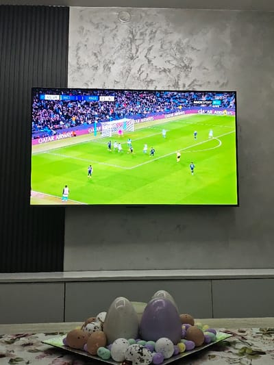 This image depicts an indoor scene within a room in Opaka, Bulgaria. A large, wall-mounted flat-screen television dominates the upper two-thirds of the frame, displaying a live soccer match. The screen shows a green soccer field with multiple players in light blue and dark uniforms. The scoreboard indicates "MCI 1 1 RMA" with a time of "45:59" and an additional timer of "11:41". A "MAXSPORT LIVE" logo is visible in the upper right. The background of the television display shows a stadium crowd.

Below the television, a light grey, low-profile cabinet or shelf unit is visible. The wall behind the television features two distinct sections: a dark, vertically ribbed panel on the left and a light grey, mottled, textured surface on the right. A small, circular, light-colored object, possibly a smoke detector or light, is affixed to the ceiling above the textured wall.

In the foreground, on a table covered with a floral-patterned tablecloth, rests a decorative centerpiece. This centerpiece consists of several large, smooth, egg-shaped objects, including one off-white and one light purple. These are surrounded by numerous smaller, speckled eggs in various shades of brown and white with black spots, along with small, pastel-colored candies or tiny eggs in light purple, yellow, and light green. The centerpiece is contained within a silver-colored, rectangular tray.