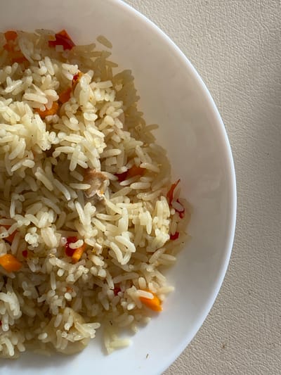 A close-up, overhead shot shows a white bowl filled with rice and diced vegetables, including carrots and possibly red bell peppers. The rice appears to be cooked and fluffy, mixed with small pieces of what might be chicken. The bowl is placed on a textured, light-colored surface, possibly a table. There is no indication of the city of Ostrovki, Russia.