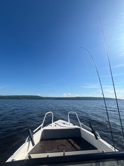The image shows the bow of a white motorboat on a dark blue body of water. Two fishing rods are propped up on the right side of the boat, with fishing lines extending into the water. The sky is a bright, clear blue with a few wispy clouds and the sun is shining, casting a glare on the right side of the image. In the distance, a tree-covered shoreline stretches across the horizon. The location is identified as Siukterka, Russia.