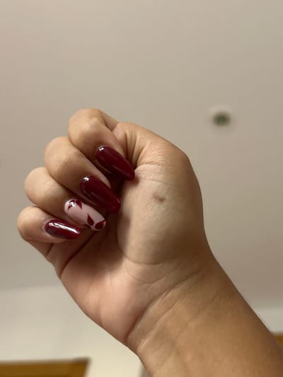 The image shows a close-up of a person's left hand with dark red, coffin-shaped acrylic nails. Four of the nails are a solid dark red color. The index finger nail is a lighter pink with dark red leaf-like shapes painted on it. The hand is partially clenched into a fist. The background appears to be a plain, off-white wall with a faint light fixture visible towards the top. There is no indication of the city of Pueblo Salinas, Spain, or any specific environmental details that would suggest its location. The scene is indoors.