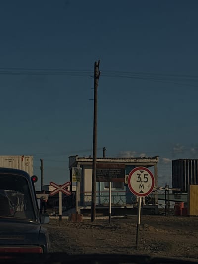 The image depicts a scene at a railway crossing in Töretam, Kazakhstan. The perspective is from inside a vehicle looking out, with the image rotated 90 degrees counter-clockwise.

The central subject is a tall wooden utility pole with wires strung across it, stretching into the clear, deep blue sky. To the right of the pole, a white and red circular sign indicates a height restriction of 3.5 meters. Below this sign, a yellow triangular warning sign with an "X" symbol is visible. A red and white "X" shaped sign, commonly associated with railway crossings, is also present.

In the background, there are several structures. A weathered building with a blue facade and a corrugated metal roof is prominent. Further in the distance, what appears to be a multi-story building with dark, horizontal lines suggests modern architecture, possibly an apartment block or office building. A white shipping container is partially visible on the left side of the frame.

The ground is a rough, gravelly texture, characteristic of unpaved areas often found near infrastructure like railway lines. The lighting suggests it is daytime, with bright sunlight casting shadows. There are no people visible in the image. The overall atmosphere is one of everyday infrastructure in a remote or developing area. The text on the signs is difficult to read due to distance and image quality, but the numerical restriction and the "X" symbols are discernible.