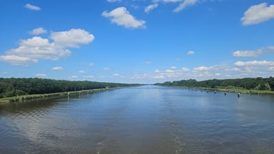The image shows a wide, calm canal under a bright blue sky with scattered white clouds. On both sides of the canal, there is a dense line of green trees. Along the banks of the canal, there are rows of short, white-capped posts. In the distance, a small boat is visible on the water. The scene appears to be a tranquil waterway in Kudensee, Germany.