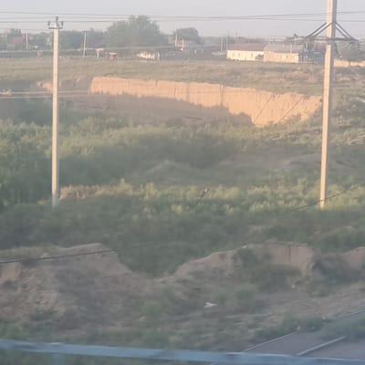 The image captures a landscape near the city of Arys, Kazakhstan. In the foreground, a blurred metal barrier is visible at the bottom. Behind it, a sloped, earthy embankment covered in sparse vegetation dominates the lower half of the image. A distinct, lighter-colored, eroded cliff face with horizontal striations is visible in the middle ground, suggesting a change in terrain or a man-made excavation. Further back, more vegetation, primarily green shrubs and trees, covers the undulating landscape. In the distance, several buildings, including houses with red and light-colored roofs, are scattered across the horizon. Two utility poles with wires are prominent features in the scene, one in the center and another to the right, indicating the presence of infrastructure. The sky is a pale, hazy blue, suggesting a bright but possibly overcast or dusty day. The overall impression is a rural or semi-rural setting with evidence of both natural and human influence on the environment.