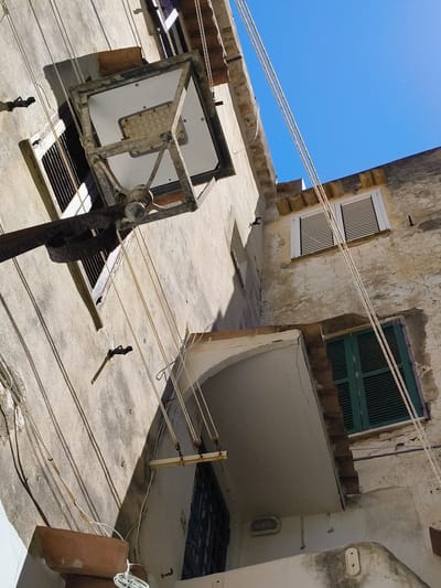 An upward-angle, exterior, close-up shot features a weathered, pale beige stucco building in Sperlonga, Italy. The bright blue sky is visible in the upper right corner. A large, metal street lamp with a white panel is suspended from a dark pole on the left side of the frame, angled diagonally. The lamp fixture is made of weathered, gray metal with visible bolts. Multiple white electrical cords run from the lamp, some attached to the building's facade, and others suspended in the air.

The building has multiple windows. On the upper left, a window is partially visible with a metal grate. To the right of this, a second window is covered by white slatted blinds. Below this, a darker window with a metal grate is visible. Further to the right and lower, a window is covered by dark green slatted shutters.

The stucco walls show signs of age and weathering, with cracks and patches of discoloration. Shadows are cast on the wall from the lamp and possibly other unseen objects. A small, brown, planter-like object is positioned at the bottom left of the frame, with white electrical cord wrapped around it. A small, white, oval light fixture is attached to the lower right portion of the building. The overall scene suggests an older urban environment.