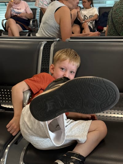 The image shows a young boy sitting in an airport waiting area, with his leg and shoe prominently displayed in the foreground. The boy is wearing an orange t-shirt and light gray shorts. He is looking towards the camera with a mischievous expression. In the background, other people are seated, appearing to be waiting for a flight. The seating appears to be typical airport chairs. The environment suggests an indoor public space, likely an airport terminal. The image was captured in the city of Bol'shie Kabany, Russia.