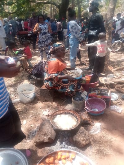 The scene depicts an outdoor gathering in Kagoma, Nigeria, characterized by food preparation and a crowd of people under a canopy of trees. In the mid-foreground, a woman, clad in an orange top, patterned skirt, and headwrap, is seated on a low dark block. She holds a white bucket filled with light-colored, diced food. Directly in front of her, a dark frying pan containing bubbling oil and cooking light-colored items rests on three large stones over an open fire with visible embers. Surrounding this cooking area are various containers including a white plastic bucket with liquid, a black pot with dark contents, a pink plastic colander, and a silver tray holding fried golden-brown food items (possibly plantains or yams) at the bottom of the frame.
Numerous individuals are present in the mid-ground and background. A person in a blue and white striped shirt is partially visible in the immediate foreground, handling a piece of light-colored food. Behind the seated woman, a woman in a black and white patterned dress with an orange jacket stands. Further back, a woman in a light patterned top and ripped blue jeans stands, while a man in a dark shirt and pants stands next to a small child in a white shirt. Two other young children are seated on the ground to the left. The background reveals a dense congregation of adults and children, with at least one motorcycle visible. The ground is unpaved earth, consistent with an outdoor setting under trees providing shade, and the lighting indicates daytime.