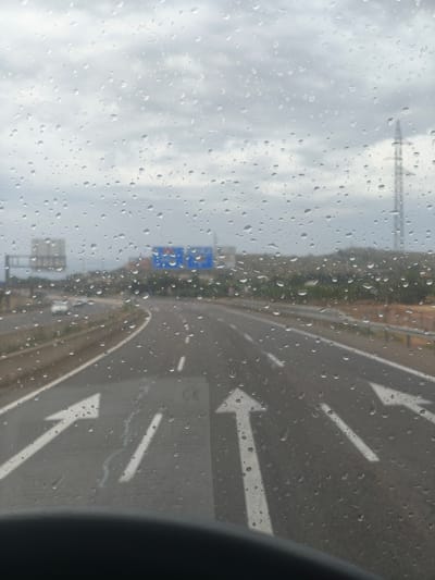 The image depicts a view through a rain-streaked window, likely that of a vehicle. The scene outside is a highway with multiple lanes. Several white directional arrows are painted on the road surface, indicating the direction of travel. In the distance, there are road signs, some of which are blue with white text. A power pylon is visible against a cloudy sky. The surrounding landscape appears to be dry and hilly, with some vegetation. The overall impression is one of overcast weather and travel on a highway. The location is identified as Llíria, Spain.
