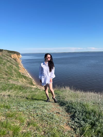 A woman with long dark hair is standing on a grassy cliff, wearing a white oversized shirt and sandals. She is facing the camera with her head slightly tilted. Behind her, there is a wide expanse of water under a bright blue sky. The cliff is covered in grass and some low-lying plants. In the distance, the opposite bank of the water body is visible. The location is Шуран, Russia.