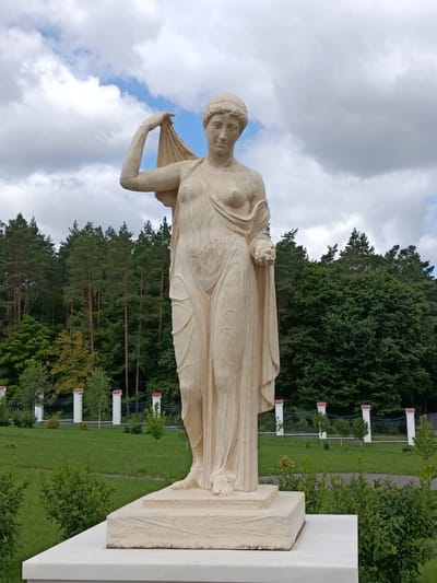 A full shot captured from a low angle shows a light-colored stone sculpture of a woman in a draped robe standing on a pedestal. The sculpture is centrally located in the frame, with a blurred background of green trees and a cloudy sky. The woman is depicted with her left arm raised, holding a piece of fabric, and her right hand is lowered, holding what appears to be a fruit. The sculpture is positioned on a multi-layered pedestal, with the lowest layer being a white marble slab. In the background, a manicured lawn with scattered bushes and a fence with decorative white posts and red caps is visible. Beyond the lawn and fence, a dense forest of pine and deciduous trees stretches across the horizon, with a partly cloudy sky above. The setting appears to be an outdoor park or garden. The sculpture is a representation of a classical figure, possibly a goddess or nymph. The overall scene evokes a sense of tranquility and artistic appreciation. The sculpture is located at the Kosava Castle in Kosava, Belarus.