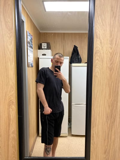 A male individual is depicted in a mirror selfie taken indoors in Марфино, Russia. The man, with short buzzed hair and light facial stubble, wears a black short-sleeved t-shirt and black athletic shorts with a grey stripe on the side. A dark-colored tattoo, resembling a stylized skull or monstrous face, is visible on his left lower shin. He holds a black-cased smartphone in his right hand, angled downwards, capturing his reflection.
The environment reflected in the mirror features light-colored wood paneling on the walls. A rectangular white LED light fixture is mounted on the ceiling. On the left wall, a multi-page calendar is affixed. Two white upright refrigerators are present; a brown cardboard box rests on the top of the left refrigerator, and a black drawstring backpack is on top of the right refrigerator. The floor appears to be a light beige or tan material. The mirror itself has a dark, thin frame and occupies the majority of the foreground.
