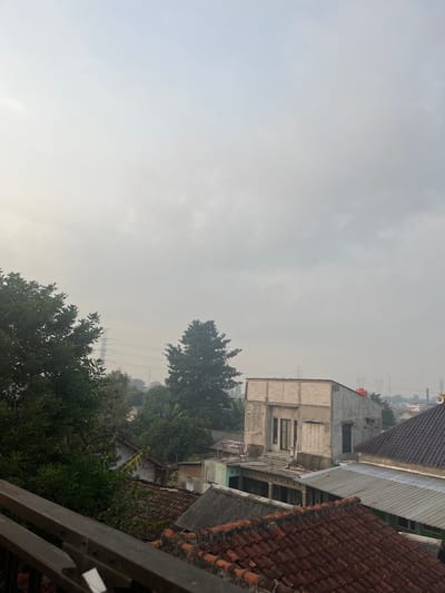 The image captures an overcast sky over a densely populated urban area. In the foreground, a metal railing overlooks a cluster of rooftops, primarily made of terracotta tiles and corrugated metal. Beyond these, various buildings are visible, some appearing partially constructed with exposed brickwork, while others are complete with windows and doors. Trees with dense green foliage are interspersed throughout the scene, particularly on the left side and towards the middle, obscuring some of the structures. In the background, power lines stretch across the hazy sky, supported by tall transmission towers, indicating the presence of electrical infrastructure. The overall impression is one of a residential neighborhood under a muted, possibly polluted, sky. The location is identified as Pasir Gadung, Indonesia.
