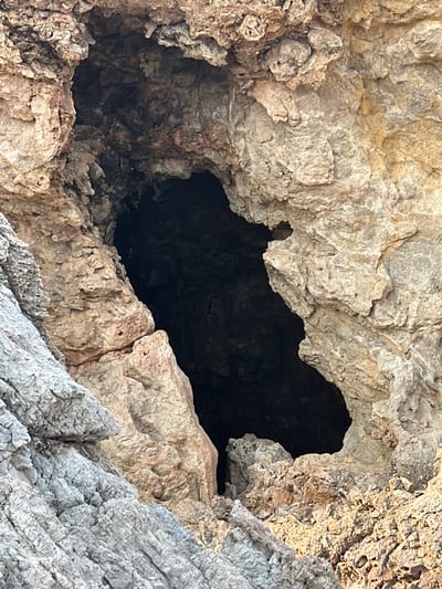 An image captured in Font De Sa Cala, Spain, depicts the entrance to a natural rock grotto or sea cave. The grotto entrance, centrally located, is an irregular, dark aperture carved into a large, textured rock formation. The rock surrounding the entrance displays varied coloration, predominantly shades of grey, tan, and reddish-brown, indicative of limestone or similar sedimentary rock, with rough, weathered surfaces. The interior of the grotto is entirely dark, suggesting significant depth or lack of internal illumination. The lower left quadrant of the frame shows a distinct slope of lighter grey, horizontally stratified rock leading towards the grotto. No individuals, man-made objects, or visible actions or interactions are present within the scene. The lighting suggests an outdoor environment, with direct illumination on the exterior rock surfaces, while the grotto interior remains shadowed.
