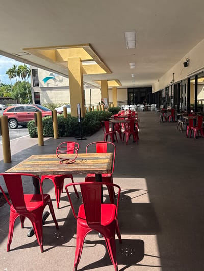 An outdoor commercial dining patio is depicted under a covered walkway, situated within Aventura ParkSquare in Aventura, United States, during daylight hours.
In the immediate foreground, a rectangular table with a distressed wooden top and a red metal spiral centerpiece is encircled by four empty red metal chairs, casting distinct shadows on the paved ground. Similar sets of red metal tables and chairs extend into the midground along the walkway, some fully illuminated by sunlight, others partially shaded. To the left, a low green hedge separates the pedestrian area from a parking lot, where a red SUV is parked. Beyond the parking area, palm trees and dense foliage obscure the view of a multi-story building featuring a prominent blue and white abstract mural.
To the right, the covered walkway abuts several commercial storefronts characterized by large, dark-framed glass windows. Further into the background, additional white tables and chairs are visible along the continued walkway. The area appears unoccupied; no individuals are visible seated at the tables or engaging in activities. The overhead ceiling of the walkway is light-colored and equipped with recessed rectangular lighting fixtures.
