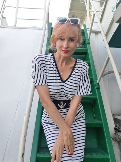 A woman with pinkish-blonde hair, wearing white sunglasses, a navy and white striped V-neck dress with a dark belt featuring an anchor emblem, and white earrings, stands on green steps. She has on red lipstick and a silver necklace. Her arms are crossed in front of her, and her hands are visible. She appears to be on the deck of a ship, suggested by the white railing and hull. The background is bright and overcast. The location is not specified to be in Shchetinino, Russia.