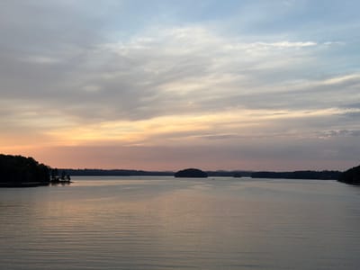 The image captures a wide-angle landscape of Lake Lanier near Buford, Georgia, United States, at dusk. The scene is divided horizontally by the lake and the sky. The upper sky displays light blue and gray tones with scattered, diffused clouds, transitioning downwards to horizontal bands of orange, pink, and light purple near the horizon, indicative of sunset. The calm water surface, occupying the lower half of the frame, exhibits subtle, uniform ripples and reflects the sky's muted colors. Dark, densely forested landmasses form the left and right shorelines, with several tree-covered islands positioned within the lake in the mid-distance. A prominent wooded point extends from the left. Distant, indistinct landforms are visible along the far horizon. No human figures are present. Several small, dark specks, possibly watercraft, are faintly visible on the distant water.