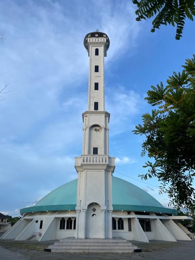 The image depicts the Masjid Raya Mujahidin mosque in Pasar Paloh, Pontianak City, West Kalimantan, Indonesia. The minaret and dome of the mosque are the central focus, positioned against a partly cloudy blue sky. Green foliage is visible in the upper corners of the frame. The building is white with a bluish-green dome. The mosque appears to be in an urban setting.