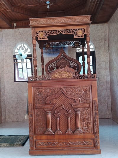 This media file captures a wooden minbar, a raised pulpit used in mosques for delivering sermons, located inside a room with tiled walls and a wooden ceiling. The minbar is elaborately carved with intricate patterns, including Arabic calligraphy and decorative motifs. The calligraphy on the minbar appears to be in gold.
The setting is a mosque or a prayer hall, suggested by the presence of the minbar and the overall architectural style, including arched windows. The light in the room appears to be artificial, with a light bulb hanging from the ceiling, and possibly natural light filtering through the windows. The windows have a decorative, arched top and are partially covered by thin curtains. A small, green prayer rug is visible on the tiled floor in front of the minbar. A ceiling fan is also present in one of the windows.
There are no people visible in the image. The scene is static, focusing on the detailed craftsmanship of the minbar. The time of day is not explicitly indicated, but the artificial lighting suggests it could be anytime, though the natural light through the windows hints at daytime. The weather is not discernible.
The location is identified as Sawang, Indonesia. The design of the minbar and the surrounding architecture may be characteristic of Indonesian mosque design. The overall impression is one of reverence and detailed artistic expression within a place of worship. The visible text is Arabic calligraphy on the minbar.