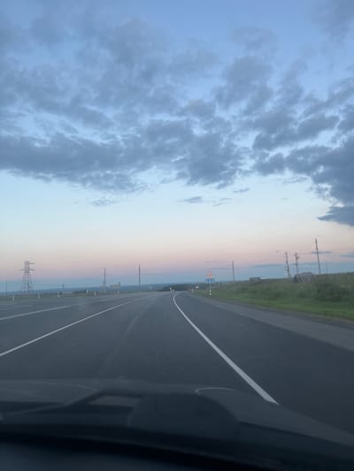 This media file shows a view from a vehicle traveling on a paved road in Tarasovo, Russia. The road appears to be a highway with a white dividing line and a shoulder. To the right of the road is a grassy embankment, and further in the distance are trees and power lines. The sky is visible, with dark clouds on the left and a gradient of light blue to pink towards the horizon. There are several electrical pylons visible along the side of the road, suggesting a rural or semi-rural setting. A road sign with a warning symbol is also visible. The time of day appears to be either sunrise or sunset, given the colors in the sky. The overall mood is calm and serene, with a sense of travel and open space.