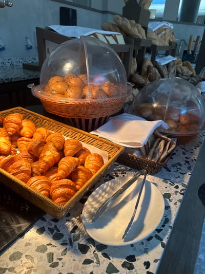 The image depicts a breakfast buffet in El Gouna, Egypt. A variety of breads, including croissants, rolls, and flatbreads, are displayed in baskets and covered containers on a speckled countertop. A white plate with tongs sits nearby. Other bread items are visible in the background on tiered racks.