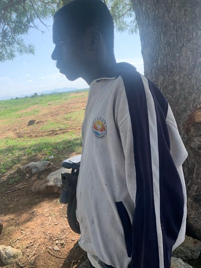 The media file shows a young person from the side, with their head tilted down. They are wearing a white jacket with dark blue stripes. A circular emblem with Chinese characters and an image of birds and a sun is visible on the jacket. The person appears to be sitting next to a tree with rough bark. In the background, there is a field with dry, brown soil and patches of green grass, suggesting a rural or agricultural setting. The sky is visible and appears bright, indicating it is daytime. A portion of a vehicle, likely a motorcycle or scooter, is visible in the lower right portion of the image, partially covered by a light-colored tarp. The person's profile is silhouetted against the bright sky, making their facial features unclear. The overall impression is of a casual outdoor scene in a rural environment in Nigeria.