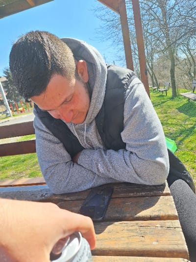 This image captures an outdoor scene in Katina, Bulgaria, on a clear, sunny day. A male individual, appearing to be a young adult, with short dark hair, is seated at a weathered wooden picnic table. He wears a grey hooded sweatshirt and a black sleeveless vest. His head is bowed, resting on his forearms, which are crossed on the table. A black mobile phone lies face-down on the table to his right. In the foreground, a hand holds a silver beverage can. The background features a grassy area with trees exhibiting sparse foliage, and several empty wooden benches. Distant structures, including a building and playground equipment, are partially visible on the far left. Wooden support beams of an overhead structure are present in the upper portion of the frame.
