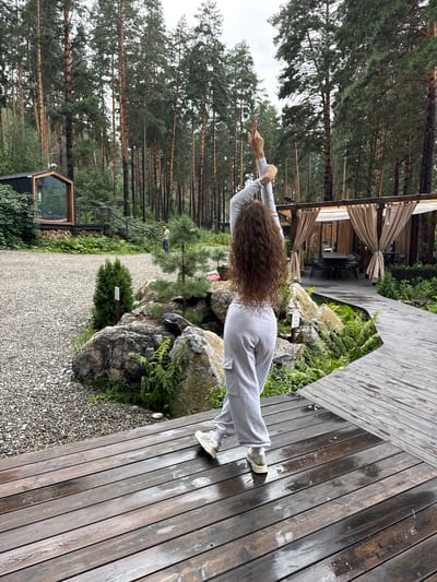 A woman with long, curly brown hair is standing on a wet wooden deck, facing away from the camera. She is wearing a light gray tracksuit and white sneakers, with her arms raised above her head, and her right index finger pointed upwards. The deck is surrounded by trees, rocks, and greenery, with a small building visible in the background. The scene appears to be an outdoor recreational area, possibly a resort or park, in Nizhnekayancha, Russia. The weather seems overcast, with the wooden deck being wet, suggesting recent rain.