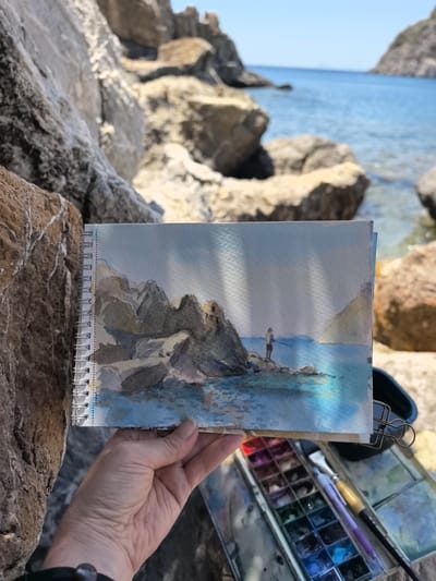 In the foreground, a person's hand holds a spiral-bound sketchbook open to a watercolor painting. The painting depicts a rocky coastline with a person fishing in the blue sea. Behind the sketchbook, a watercolor paint set and brushes are visible. The background shows the natural environment of Datça, Turkey, with large rock formations and the clear blue sea. The scene appears to be a plein-air painting session.