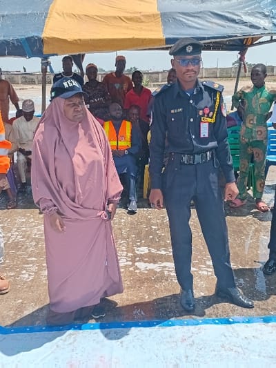 The image shows a group of people in Katabu, Nigeria (latitude: 10.71, longitude: 7.514). A woman wearing a pink abaya and a black hat is standing in front of a man wearing a navy blue uniform, a black cap, sunglasses, and a black belt. He is possibly a law enforcement officer, as he has a badge on his shirt and his uniform includes a patch with the word "LAP". There are several other people in the background, some sitting in chairs and others standing. The scene appears to be a public event. The event is taking place on a paved area. There is a large white tent in the background. The ground is wet, possibly due to rain.
