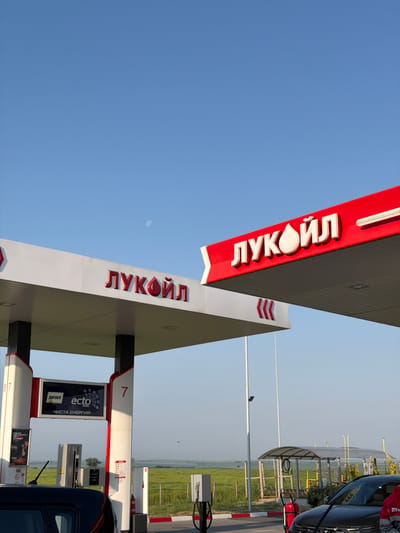 The image captures a vibrant, high-angle view of a Lukoil gas station under a clear, bright blue sky, presumably in Vodenichane, Bulgaria, given the Cyrillic script. The perspective is tilted, showing the underside and sides of two prominent red and white canopies that extend over the fueling area. Both canopies feature the distinct "ЛУКОЙЛ" (LUKOIL) logo in white, three-dimensional letters against the red stripe. The larger canopy also displays red chevron markings.
At the lower right, a black car is partially visible, parked next to gas pumps. One pump pillar clearly shows the number "7" and an advertisement for "ectо ДИЗЕЛ" (Ecto Diesel) with the tagline "ЧИСТА ЕНЕРГИЯ" (Clean Energy). Another pump, partially visible at the very bottom, shows the number "87". A red fire extinguisher is positioned near the car. The ground features red and white striped curbing, delineating the service area.
In the background, beyond the gas station structures, a simple, covered shelter (possibly a car wash or service bay) with a mesh fence is visible. Green agricultural fields stretch into the distance under the open sky, suggesting a semi-rural setting. A small, faint moon is discernible in the upper left portion of the sky, indicating it is daytime, likely morning or late afternoon. The overall weather appears sunny and calm, with no clouds. No people are explicitly visible, but their presence is implied by the active gas station environment.