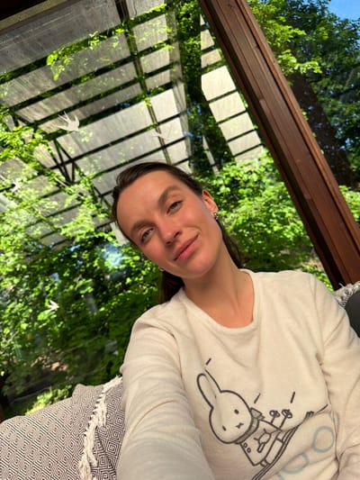 A woman wearing a white, long-sleeved shirt with a cartoon bunny is sitting inside what appears to be a sunroom or greenhouse. The room has a glass or translucent roof and walls, with a wooden frame. Outside the room, there are green trees and vegetation visible through the glass. A patterned throw blanket is draped near the woman. The woman is smiling and looking at the camera. The location is Tarasovka, Russia.